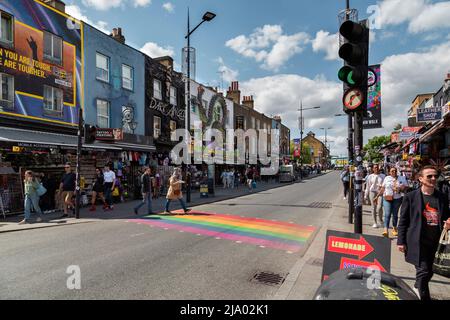 People walking across a rainbow coloured pedestrian crossing on Camden High Street. Colourful ornate shop fronts line the busy road. Stock Photo