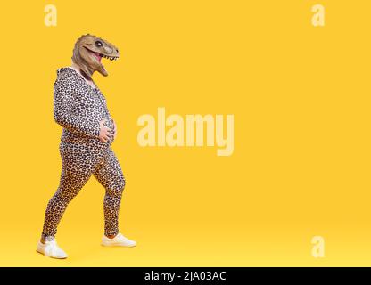 Funny fat man with big belly wearing pajamas and dinosaur mask isolated on orange background. Stock Photo