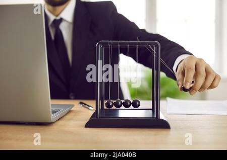Businessman sitting at office desk with laptop and playing with Newton's pendulum Stock Photo