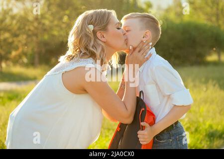 mother kissing son before going to school . stylish and young schoolboy in a white shirt and jeans with a backpack in the park Stock Photo