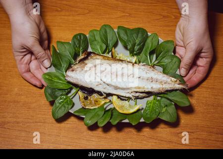 Plate, dish of oven baked white freshwater fish with lettuce leaves in woman's hands top view. Serving seafood on wooden table. Preparing food for din Stock Photo