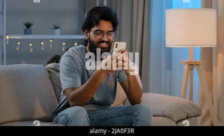 Smiling Arabian Indian man bearded male looking in smartphone smile texting with friend girlfriend messaging chatting at home sitting on couch at Stock Photo