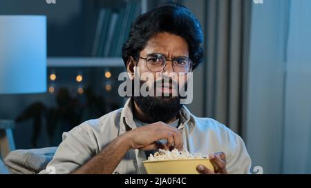 Interested attentive Arabic Hispanic Indian bearded man guy in glasses eating popcorn watching TV show series horror movie late night feeling scary Stock Photo