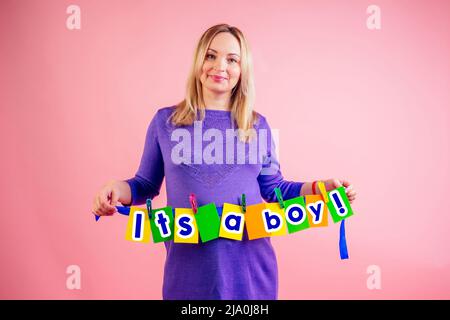 beautiful smiley pregnant big belly woman at 9 months of pregnancy hold a cards with the inscription 'it's a boy' in studio on pink background Stock Photo