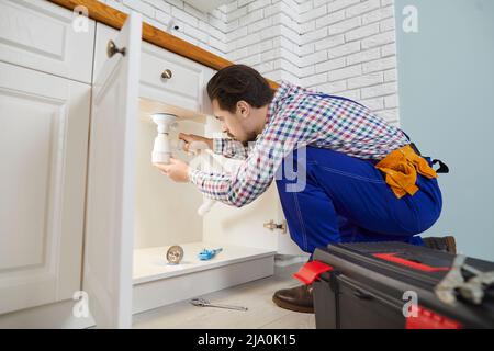 Skilled male worker plumbing repair service installs siphon under sink in kitchen in apartment. Stock Photo