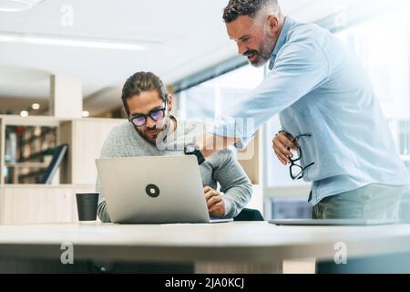 Two entrepreneurs sharing business ideas while working together in a modern co-working office. Focused businessmen using a laptop while working on a n Stock Photo