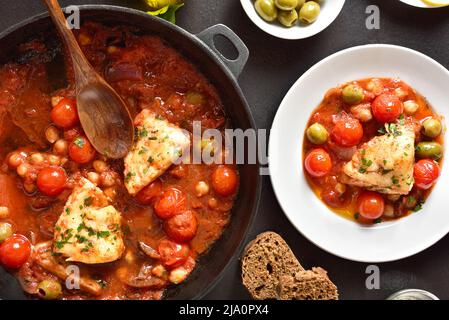 Cod stew with chickpeas, cherry tomatoes and olives over dark stone background. Top view, flat lay Stock Photo
