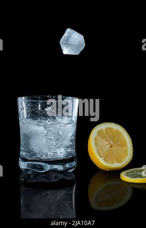 Ice cubes falling into a cocktail glass with lemon slice and reflections on a black background. Stock Photo