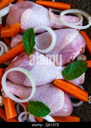 Raw chicken drumsticks with carrots, onions and green herbs in frying pan top view. Fresh meat vegetables salad spices cooking. Homemade healthy yummy Stock Photo