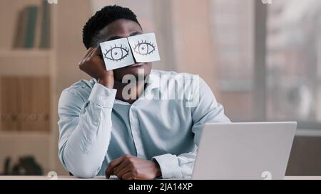 Sleepy funny african american office manager business man sitting at home workplace with sticky notes on eyes comic young tired worker has sleeping Stock Photo