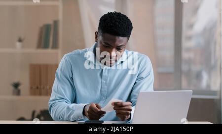 Young African focused man bank employee bookkeeper hold payment bill manage personal budget use calculator calculates expenses costs credit balance Stock Photo