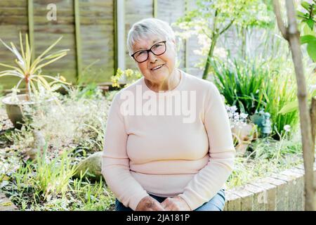 Portrait of smiling senior, mature retired grandmother in eyeglasses. Happy elderly woman smiling sitting in her green garden and enjoying retirement Stock Photo