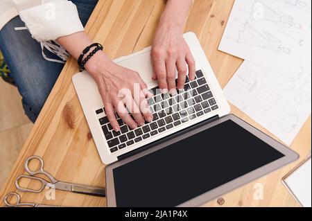 View from above to female hands on laptop keyboard on a wooden table near sketches and tailoring scissors. Copy space for advertising text on the blan Stock Photo