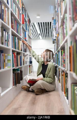 Attractive woman teacher sits on floor in school library, prepares to study with students at lesson. Stock Photo