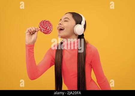 happy kid singer listen music in headphones singing with lollypop on yellow background, singer Stock Photo