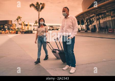 A couple of friends in protective masks chatting while leaving the airport with their luggage Stock Photo