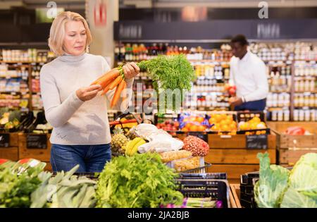 Focused middle aged woman shopping in organic food store, choosing fresh vegetables Stock Photo