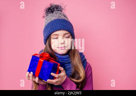 Happy little smiling blonde curly hairstyle girl in knitted blue gray hat and scarf with christmas gift box pink background studio. new Year present Stock Photo