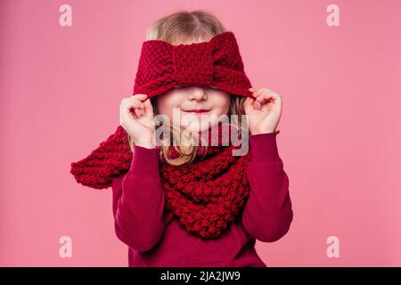 Beautiful little girl in winter warm colorful red hat and scarf dreaming Christmas night playing hide and seek in studio on pink background.portrait Stock Photo