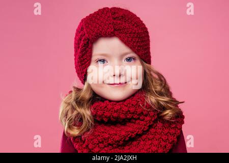 Beautiful little girl in winter warm colorful red hat and scarf dreaming thought about the gift Christmas night in the studio on a pink background Stock Photo
