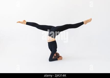 Woman in black sportswear practicing yoga doing shirshasana exercises with hanumanasana, headstand Stock Photo