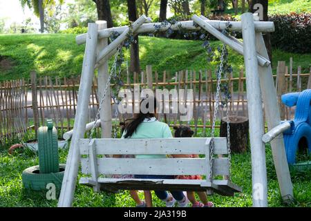 Rear view of little girls sitting on a swing under tree with mother in the summer park. Happy family having fun in the park. Stock Photo