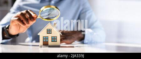 Close-up Of A Businessman's Hand Holding Magnifying Glass Over House Model Over Desk Stock Photo