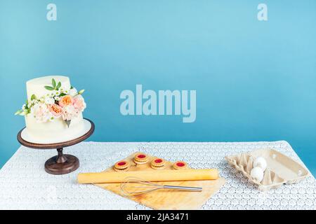 copyspace mockup working space baker confectioner pastry chef creamy white two-tiered wedding birthday cake with fresh flowers on table and cookies in Stock Photo