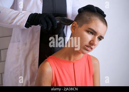 Young woman is forcibly shaving her head with an electric razor, holding typewriter in black gloves, a girl with a sad look with tears in her eyes. Close-up, copy space Stock Photo