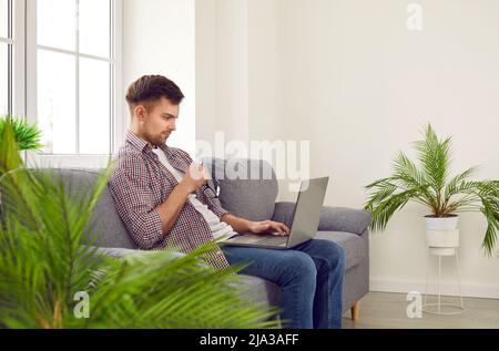 Young male freelancer with serious facial expression working remotely at home using laptop. Stock Photo