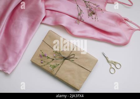 Composition with pink silk fabric, gift in craft paper and threads and scissors on white background. Flat lay, top view. Hobby, leisure concept. Stock Photo