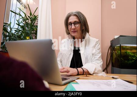 Pleasant senior Caucasian woman using laptop, works in fashion design atelier, creating clothes sketches laid on wooden desk. Creative occupation, onl Stock Photo