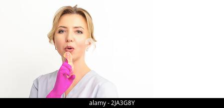 Young caucasian female beautician holding wax paste on forefinger blowing on him wearing pink gloves against a white background. Epilation concept Stock Photo