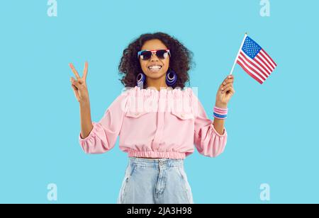 Happy dark-skinned teen girl showing victory sign holding america flag on light blue background. Stock Photo