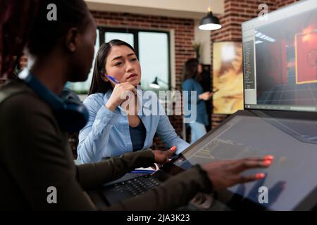 Multiethnic creative game creators sitting at desk with multiple displays while working with 3D assets. Digital interactive entertainment industry workers working with CGI. Stock Photo