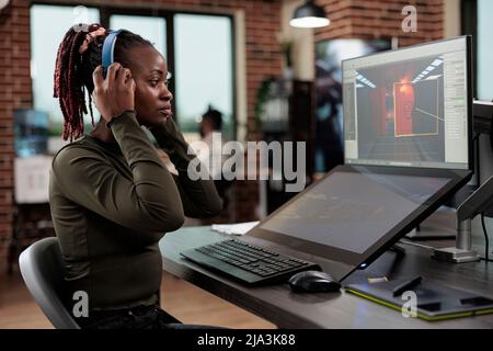 Level design department employee wearing wireless headphones while analyzing digital environment. Digital artist in office workspace sitting at desk while working on game interface. Stock Photo