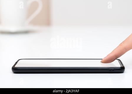 woman tapping on phone blank white screen. Smart phone on white table and female hand with finger on blank touchscreen. Woman finger presses on screen Stock Photo