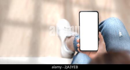 Top view mockup image of woman holding mobile phone with blank white screen. Woman in jeans sits cross-legged and holds cell phone in her hands with b Stock Photo