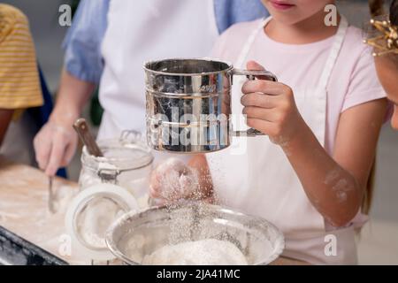 Cute little girl wearing apron passing ripe tomato to elder sister while cooking delicious salad together, kitchen interior on background Stock Photo