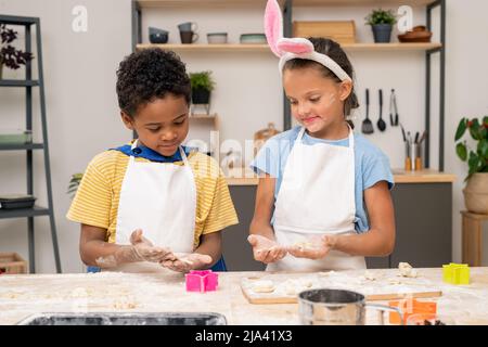 Cute blond boy rolling homemade dough on wooden table with his mom standing near by while cooking together Stock Photo