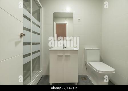 Newly renovated bathroom with a square frameless mirror, a porcelain sink with a high gloss white wood cabinet, a walk in shower and white tile Stock Photo