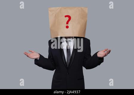 Young businessman covering his face using a craft paper bag with red drawn question mark, like a mask, for hiding his identity. Man isolated on gray w Stock Photo