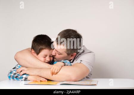 Dad hugs and kisses the son of a preschooler sitting at the table and doing his homework Stock Photo