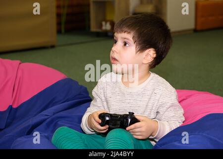 A handsome boy of 4 years old plays a game console, holds a joystick in his hands and looks away, a place for text Stock Photo