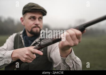 Hunter man in traditional shooting clothes on field aiming with shotgun. Stock Photo