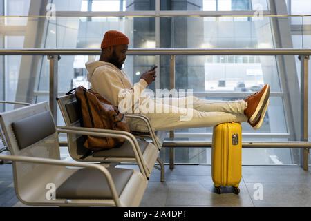 Thoughtful young hipster black man sitting in airport terminal using smartphone and waiting landing Stock Photo