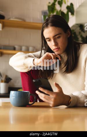 Thoughtful non-binary trans woman using smartphone in the kitchen at home Stock Photo