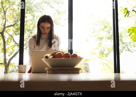 Thoughtful non-binary trans woman using laptop in the kitchen at home Stock Photo