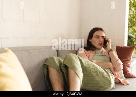 Non-binary trans woman talking on smartphone lying on the couch at home Stock Photo