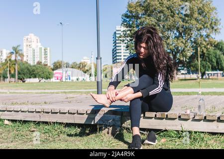 sporty young caucasian woman, sitting outdoors in a park, putting hot cream on her feet and ankle to start training in the morning. Stock Photo